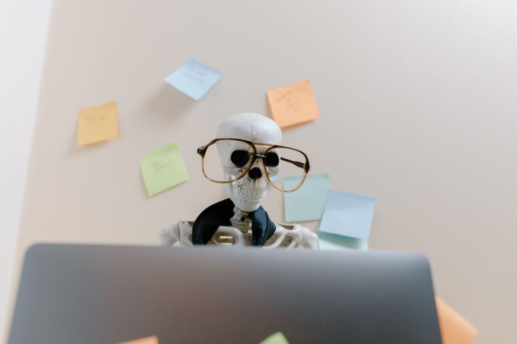 Skeleton wearing glasses uses laptop surrounded by sticky notes, symbolizing stress or burnout.